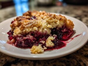 A close-up of a warm slice of mixed berry cobbler with a golden crumble topping served on a white plate.