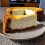 A thick slice of creamy new york cheesecake with a golden-brown graham cracker crust on a plate.
