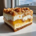 A square slice of peach cobbler cheesecake showing layers of graham cracker crust, cheesecake, peaches, and a brown sugar crumble topping.