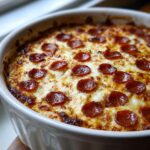 Close-up of a hot, bubbly pepperoni pizza dip topped with melted, browned cheese in a white baking dish.