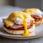Close-up of Eggs Benedict featuring a poached egg with creamy hollandaise sauce dripping down, served over ham and an English muffin.