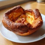 A single, perfectly risen and golden-brown yorkshire pudding sitting on a white plate.