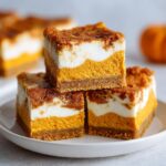 Three stacked pumpkin cheesecake bars featuring a graham cracker crust, bright orange pumpkin layer, and white cream cheese swirl.