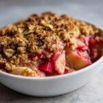 A close-up, appetizing view of warm rhubarb crisp topped with a golden oat crumble, served in a white bowl.