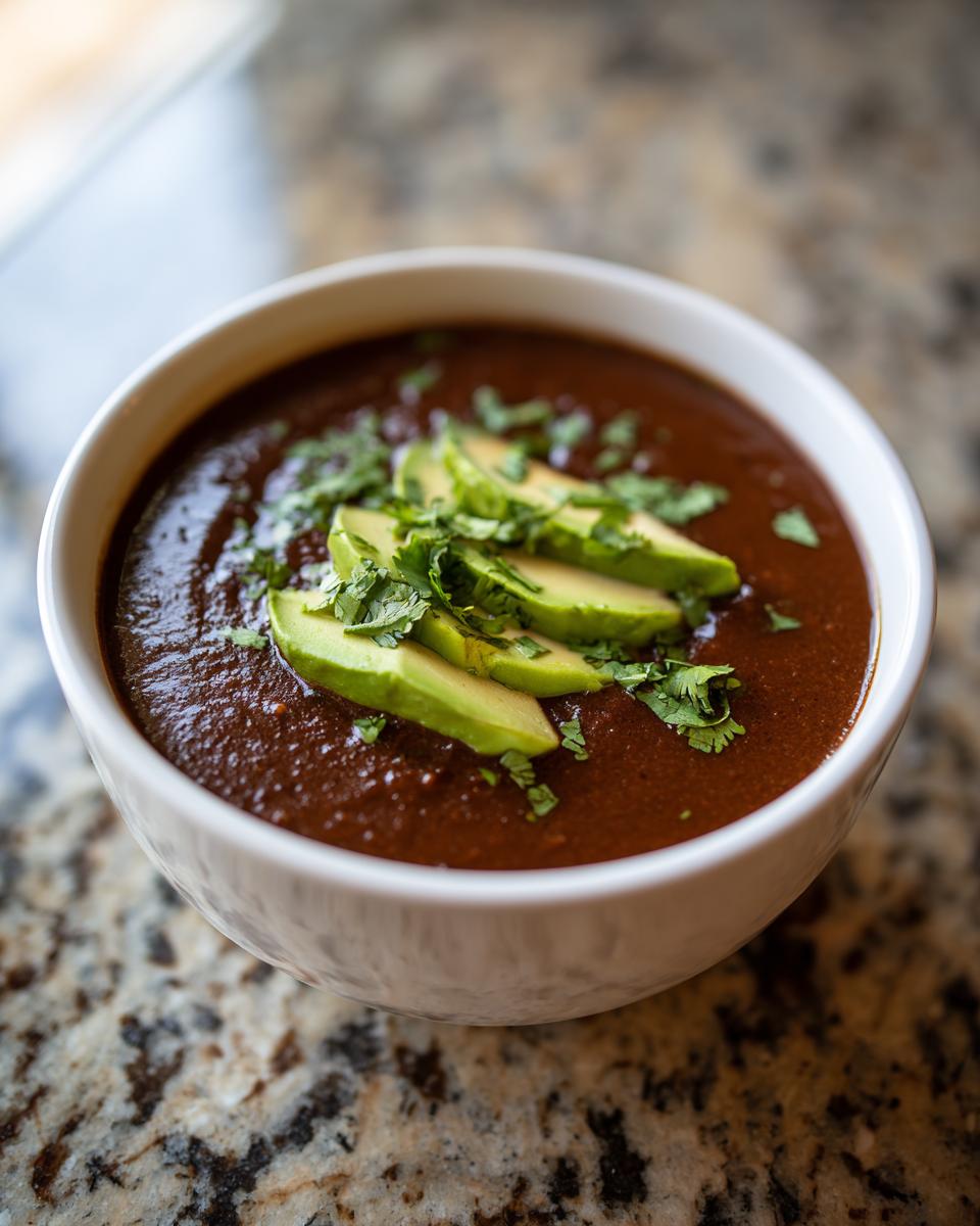 A close-up of a bowl of rich, dark black bean soup garnished with sliced avocado and fresh cilantro.
