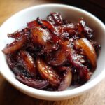 Close-up of deeply browned, glossy caramelized onions served in a small white bowl.