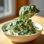 A spoonful of rich, cheesy creamed spinach recipe being lifted from a white bowl.