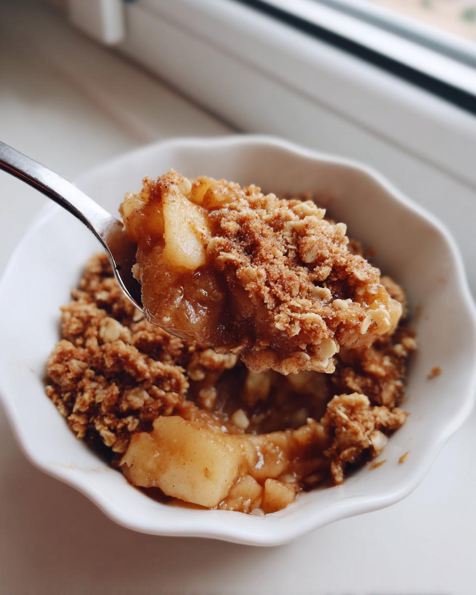 A spoonful of warm, gooey apple crisp being lifted from a white bowl, highlighting the oat topping.