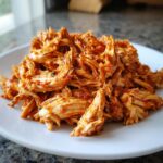 Close-up of perfectly shredded chicken tinga coated in a vibrant, savory red chipotle sauce on a white plate.