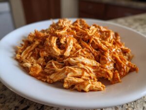 Close-up of shredded chicken tinga coated in a rich, reddish-orange sauce, served on a white plate.