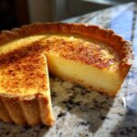 A slice cut from a silky custard pie, showing the smooth yellow filling and caramelized, sprinkled topping.