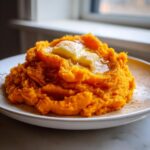 A close-up of a serving of bright orange, silky mashed sweet potatoes topped with two pats of melting butter and a sprinkle of spice.