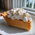 A delicious slice of rich orange butterscotch pie topped high with fluffy whipped cream, sitting on a white plate.