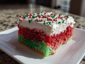 A colorful slice of christmas poke cake showing red and green layers topped with white frosting and holiday sprinkles.