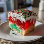 A slice of colorful christmas poke cake featuring green and red jello layers, whipped cream, and sprinkles.