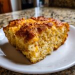 A close-up of a moist slice of cornbread casserole with visible corn kernels and a golden-brown, slightly caramelized top.