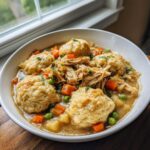 A white bowl filled with rich slow cooker chicken and dumplings, featuring shredded chicken, carrots, peas, and fluffy dumplings.