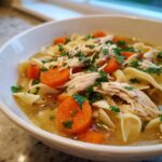 Close-up of a white bowl filled with slow cooker chicken noodle soup, featuring shredded chicken, egg noodles, and bright orange carrots.