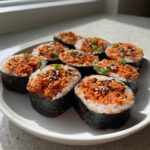A plate holding several pieces of freshly cut tuna kimbap, topped with spicy orange filling, sesame seeds, and green onions.
