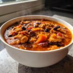 A close-up of a steaming white bowl filled with rich, thick pumpkin chili featuring ground meat, beans, and chunks of orange pumpkin.