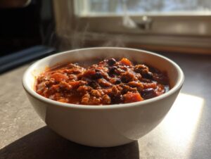 A steaming white bowl filled with rich, hearty pumpkin chili featuring ground meat and beans, set near a window.