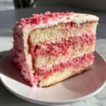 A moist slice of layered strawberry cake with white frosting and pink strawberry crumbs on top.