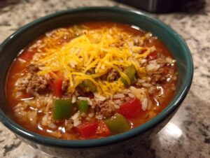 Close-up of a bowl of stuffed pepper soup featuring ground meat, rice, red and green peppers, topped with shredded cheddar cheese.