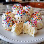 A plate of white chocolate-covered sugar cookie truffles topped with rainbow sprinkles, one cut in half showing the soft interior.