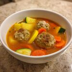 Close-up of a white bowl filled with flavorful albondigas soup featuring meatballs, carrots, zucchini, and potatoes.