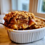 A spoonful of gooey, spiced apple crisp being lifted from a white ramekin.