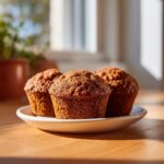 Three freshly baked, rich brown sweet potato muffins sitting on a small white plate on a wooden surface.