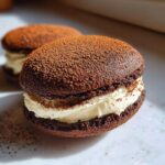 A close-up of two decadent tiramisu cookies filled with white cream and dusted heavily with cocoa powder.