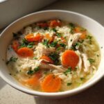 Close-up of a white bowl filled with steaming turkey soup, featuring shredded turkey, sliced carrots, rice, and fresh parsley.