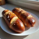 Two freshly baked pretzel dogs with a dark brown, glossy crust and coarse salt on a white plate.