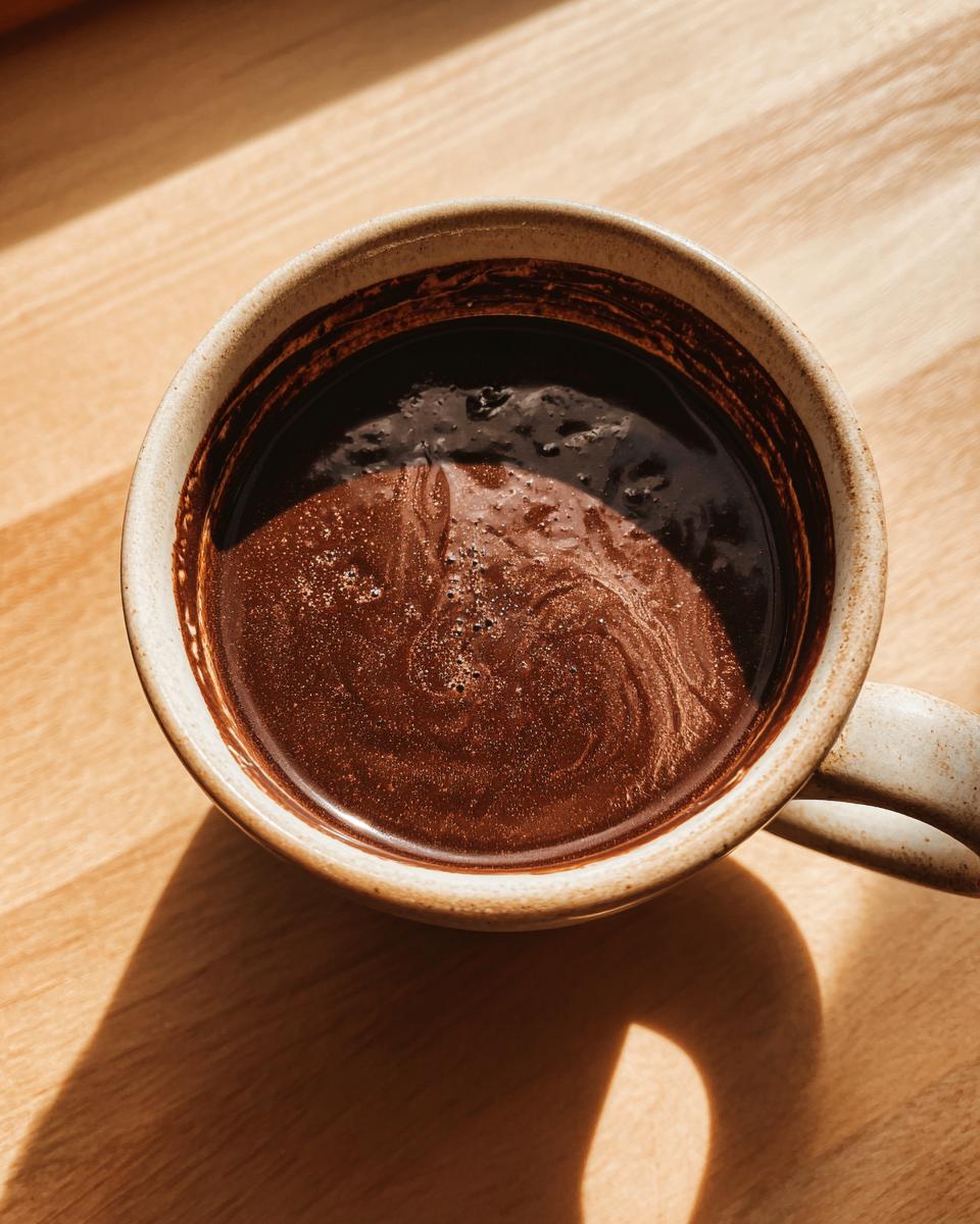 Overhead view of rich, dark french hot chocolate swirling in a rustic ceramic mug on a wooden surface.