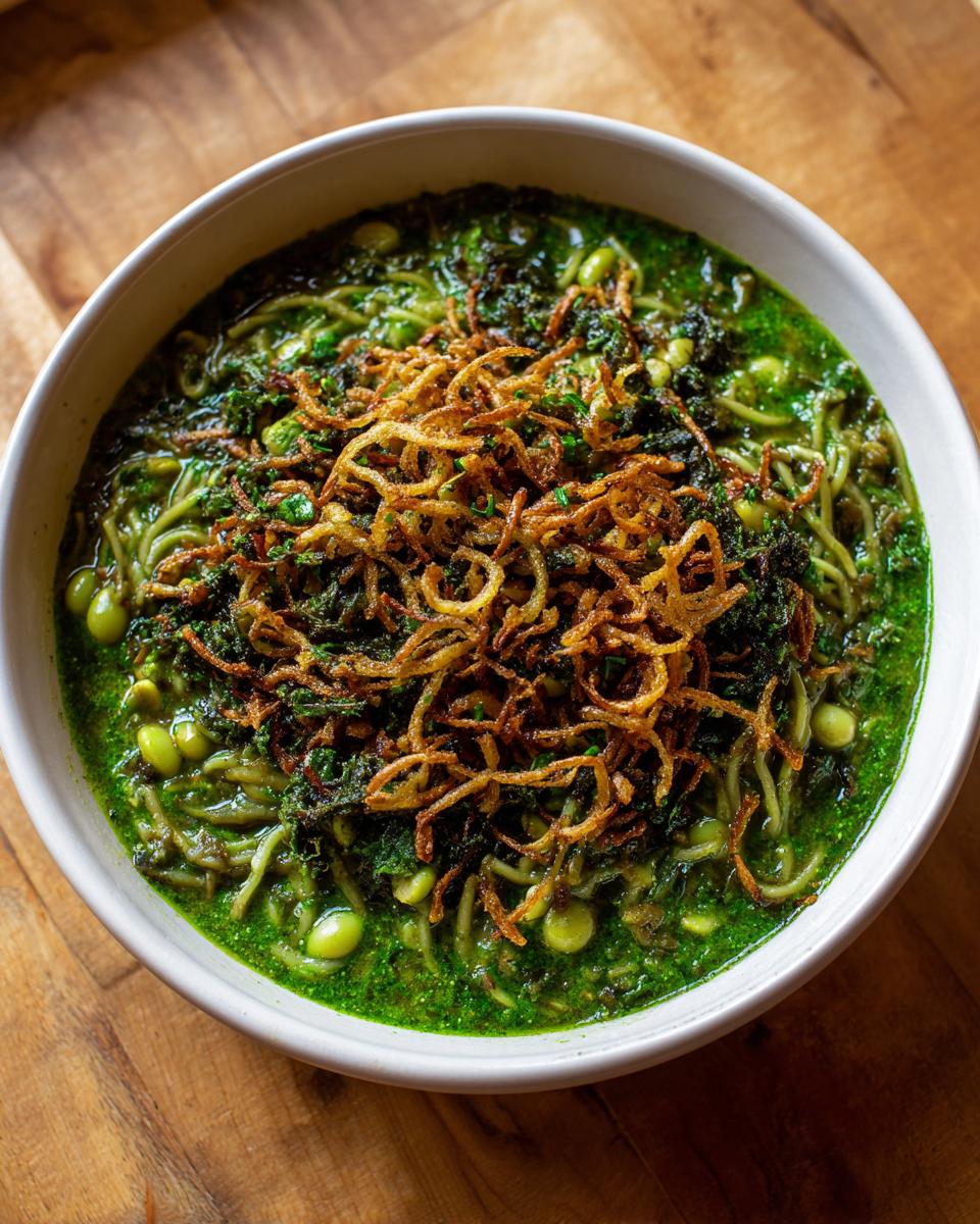 A close-up of a bowl of rich green Persian noodle soup topped with fried onions and herbs.