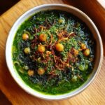 Close-up of a rich, green Persian noodle soup topped with chickpeas, fried onions, and fresh herbs.